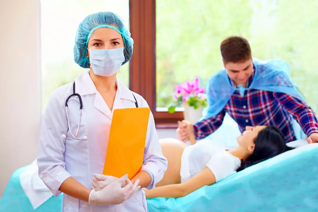 Photo of a doctor working during childbirth. In the background is a husband and wife who are waiting for the birth of their baby