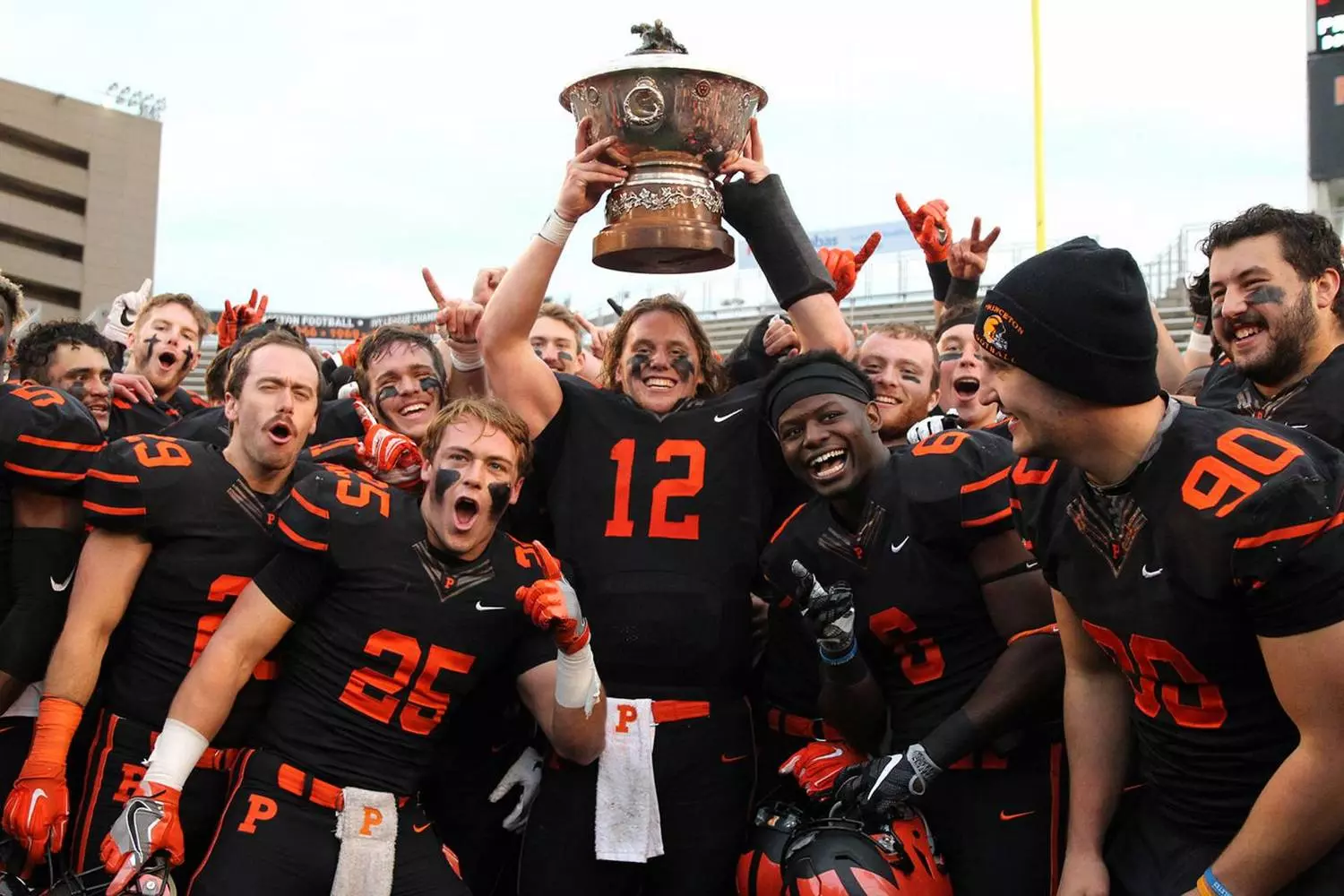 Студенты-спортсмены Принстона в форме с символикой тигра празднуют победу, улыбаясь и обнимаясь на поле