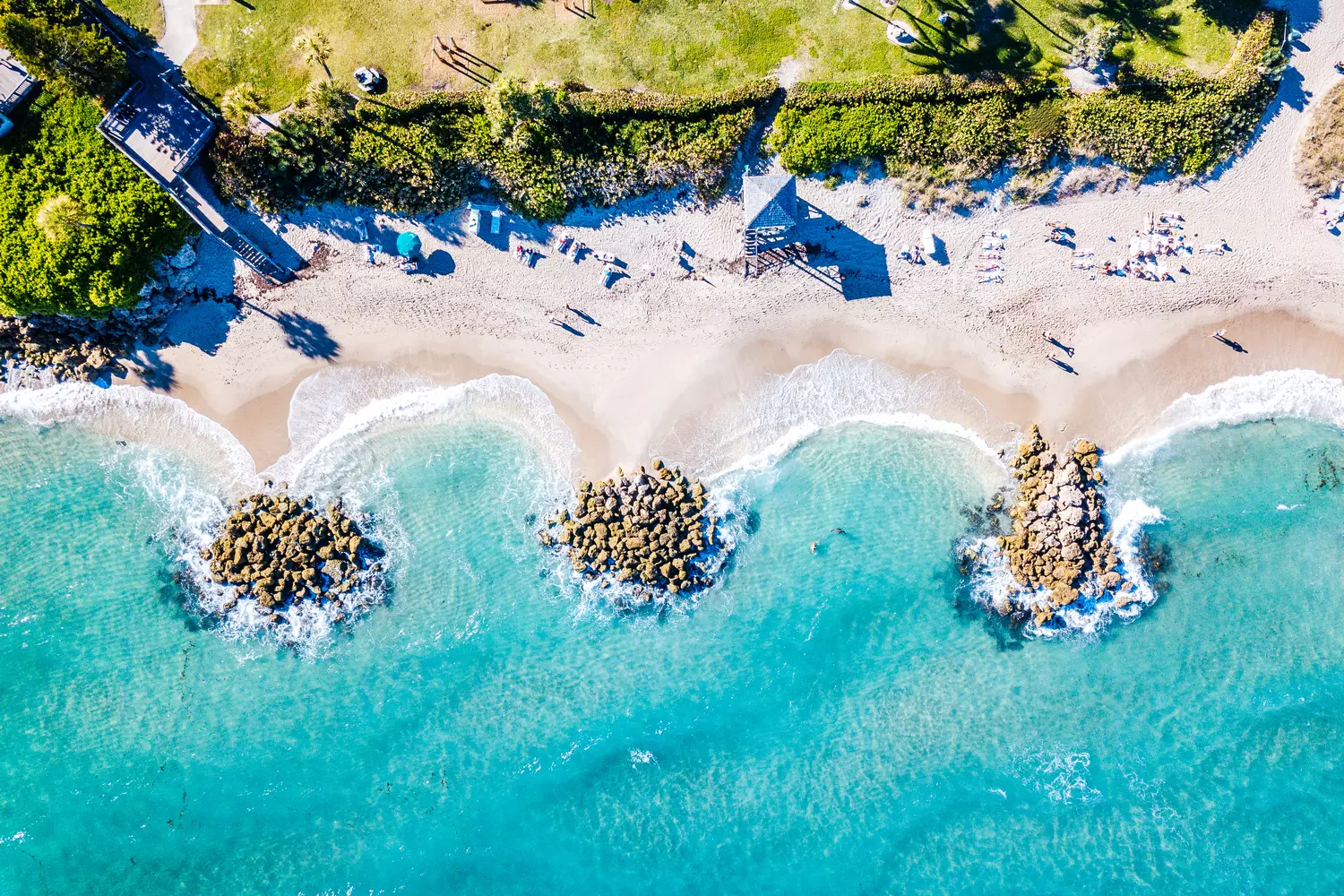 Helicopter aerial view of Fort Lauderdale beach featuring a wide stretch of golden sand, turquoise Atlantic waves crashing onto the shore, and coastal buildings seen from a bird's-eye perspective