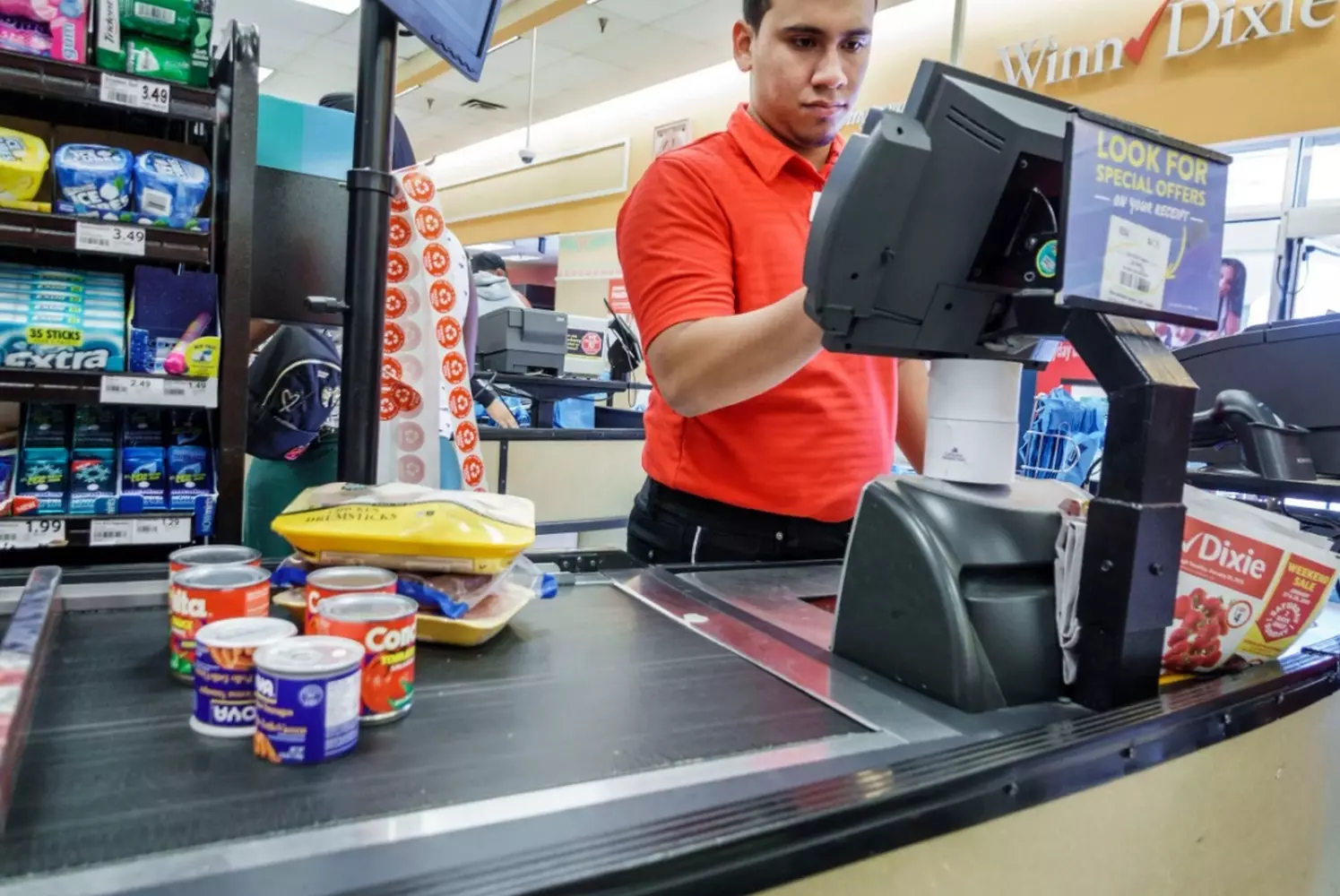 Checkout Chronicles: The American Supermarket Cashier Experience Busy checkout area in an American supermarket showing customers unloading carts onto conveyor belts, cashiers scanning items, and colorful impulse-buy displays near the registers under bright fluorescent lights