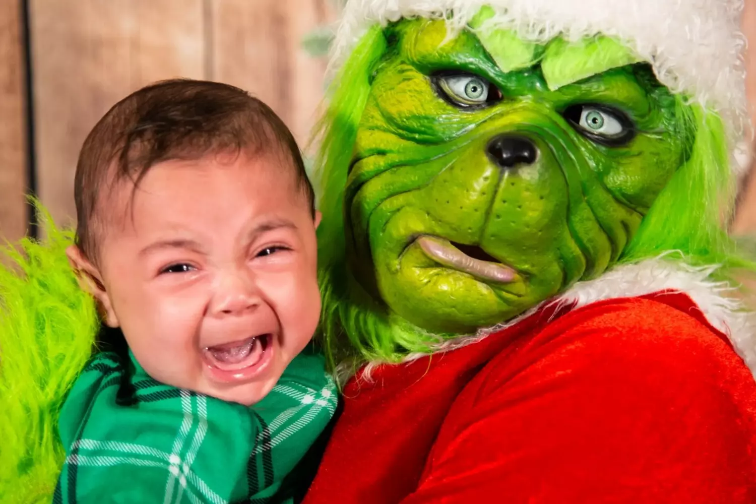 The Grinch's Prank Gone Wrong: Baby's Hilarious Christmas Terror A person in full Grinch costume with wild green fur and sinister smile leans over a terrified infant who's crying in fear, creating a darkly comedic holiday moment at a Christmas party