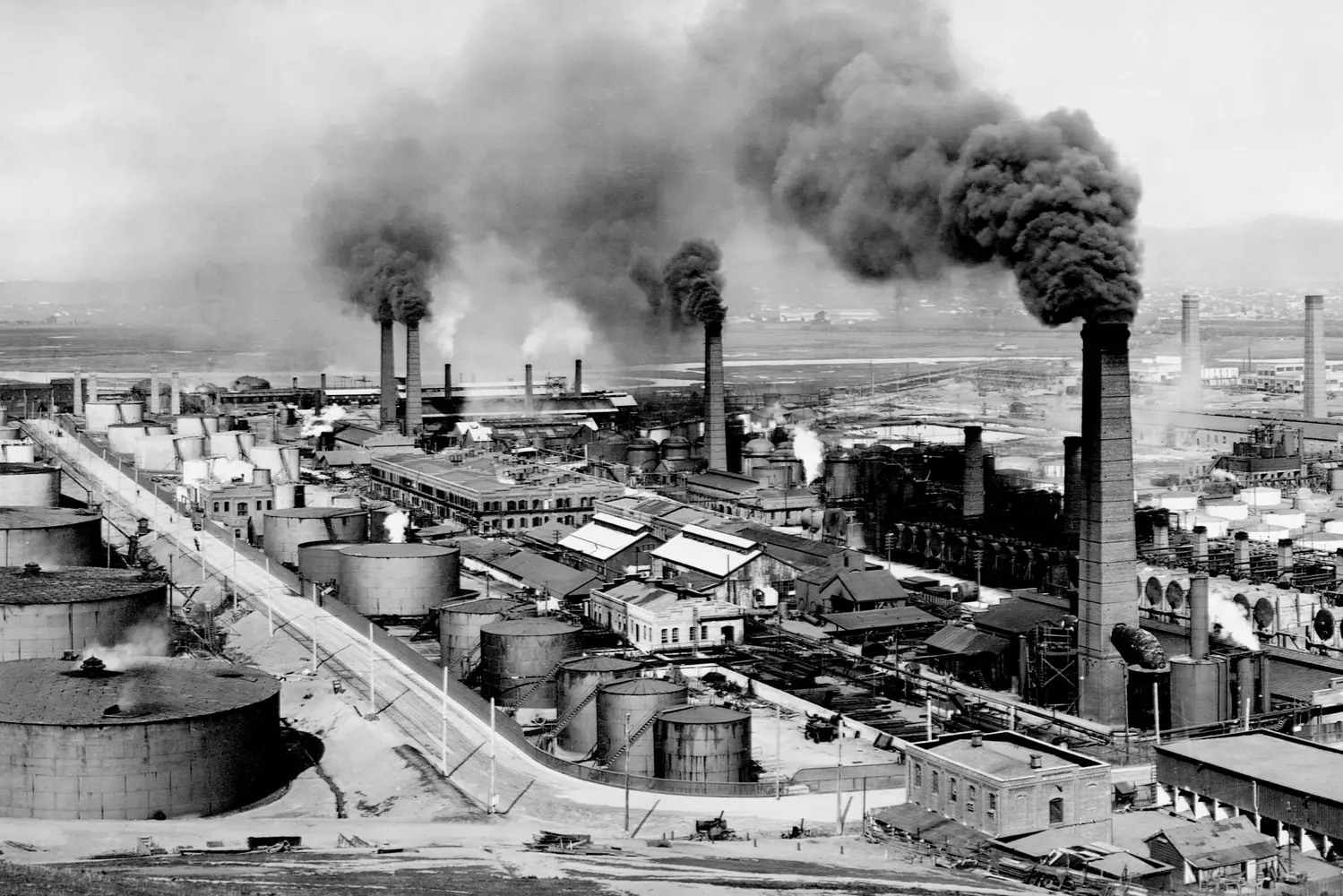 Исторический нефтеперерабатывающий завод в США: Индустриальное наследие XX века Чёрно-белое фото нефтеперерабатывающего завода в США начала XX века с дымовыми трубами, трубопроводами и резервуарами — символ зарождения нефтяной эры