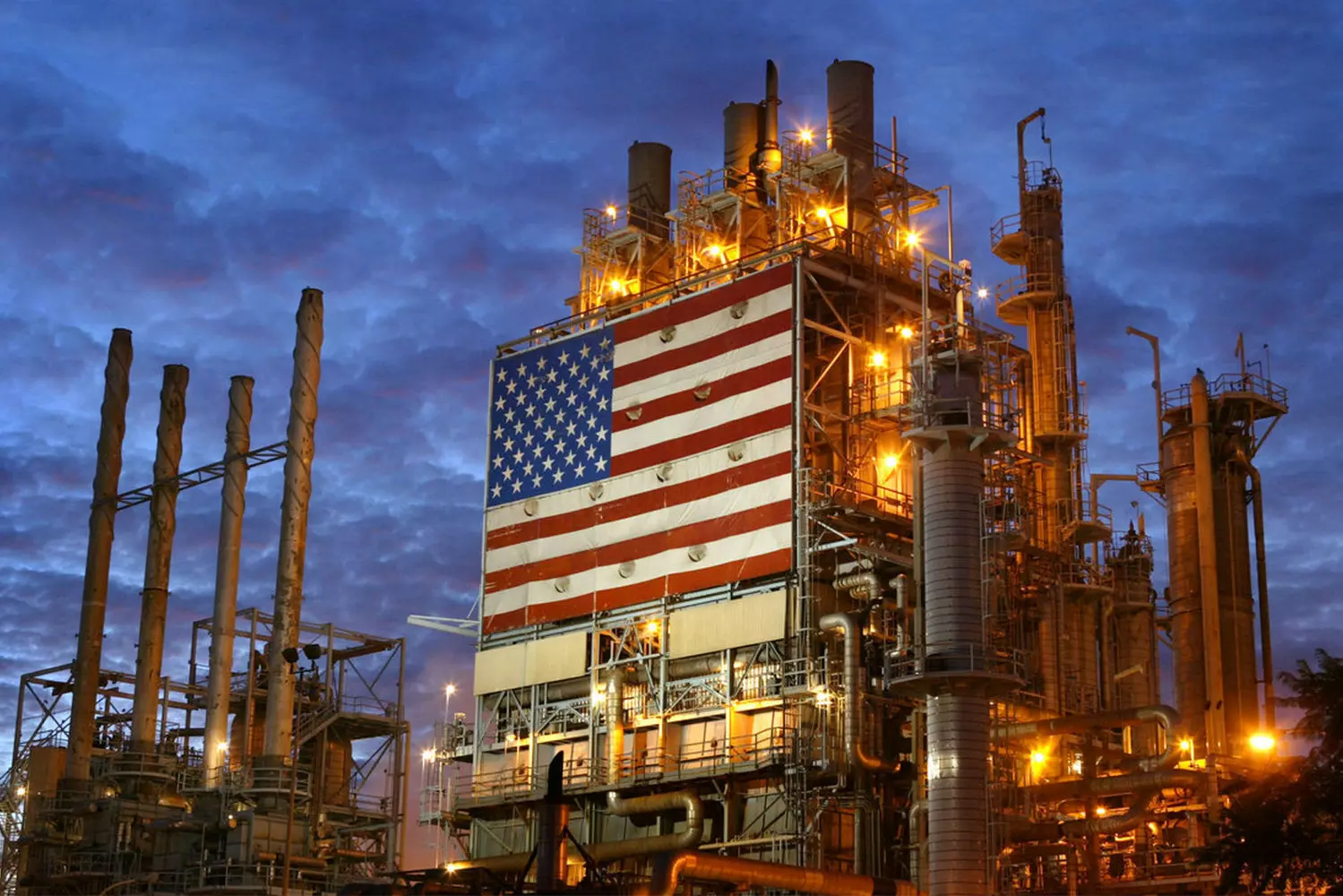 Нефтеперерабатывающий завод в США: Сердце энергетики страны Крупный нефтеперерабатывающий завод в США с дымовыми трубами, трубопроводами и резервуарами на фоне облачного неба — важный элемент американской энергосистемы