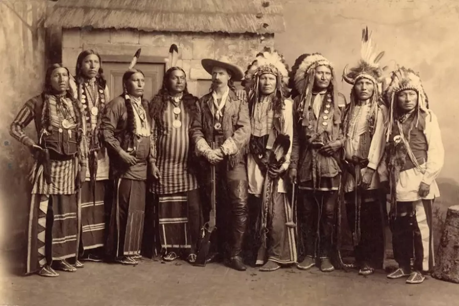 Rare Studio Portrait of a Cowboy and Native American Men A staged 19th-century studio portrait showing a white cowboy in a hat sitting alongside several Native American men in traditional clothing, posing together against a painted backdrop