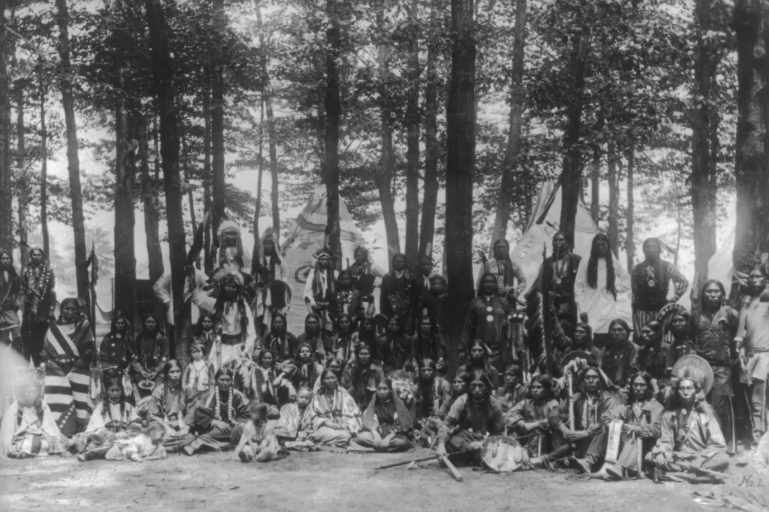 Historic Photo of a Native American Village with Tipis A black and white historical photograph of a traditional Native American village, showing multiple tipi lodges arranged near a river or on the plains, with people and horses nearby