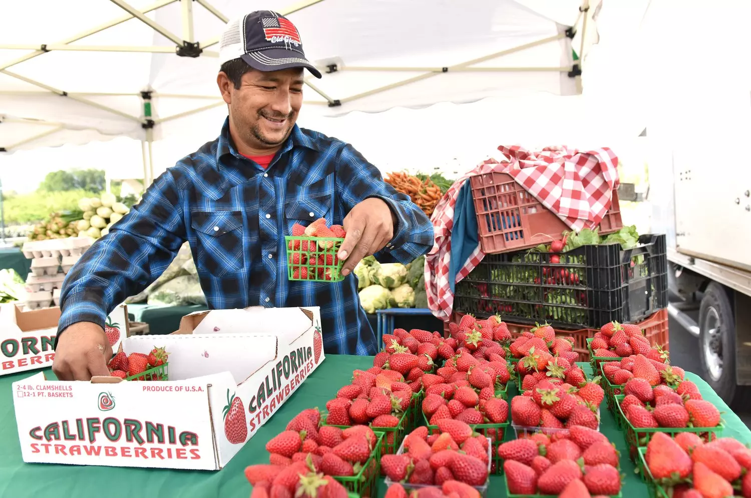 Улыбающийся продавец за деревянным прилавком выставляет корзинки с ярко-красной, спелой клубникой на открытом рынке в Америке