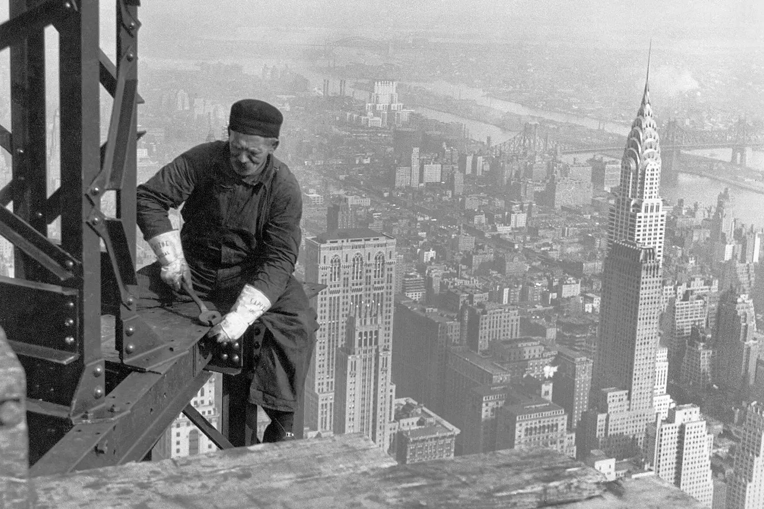 Стальные исполины: архивное фото строительства небоскрёба в США Чёрно-белое историческое фото строительства небоскрёба в США: рабочие на стальных балках на фоне городского горизонта