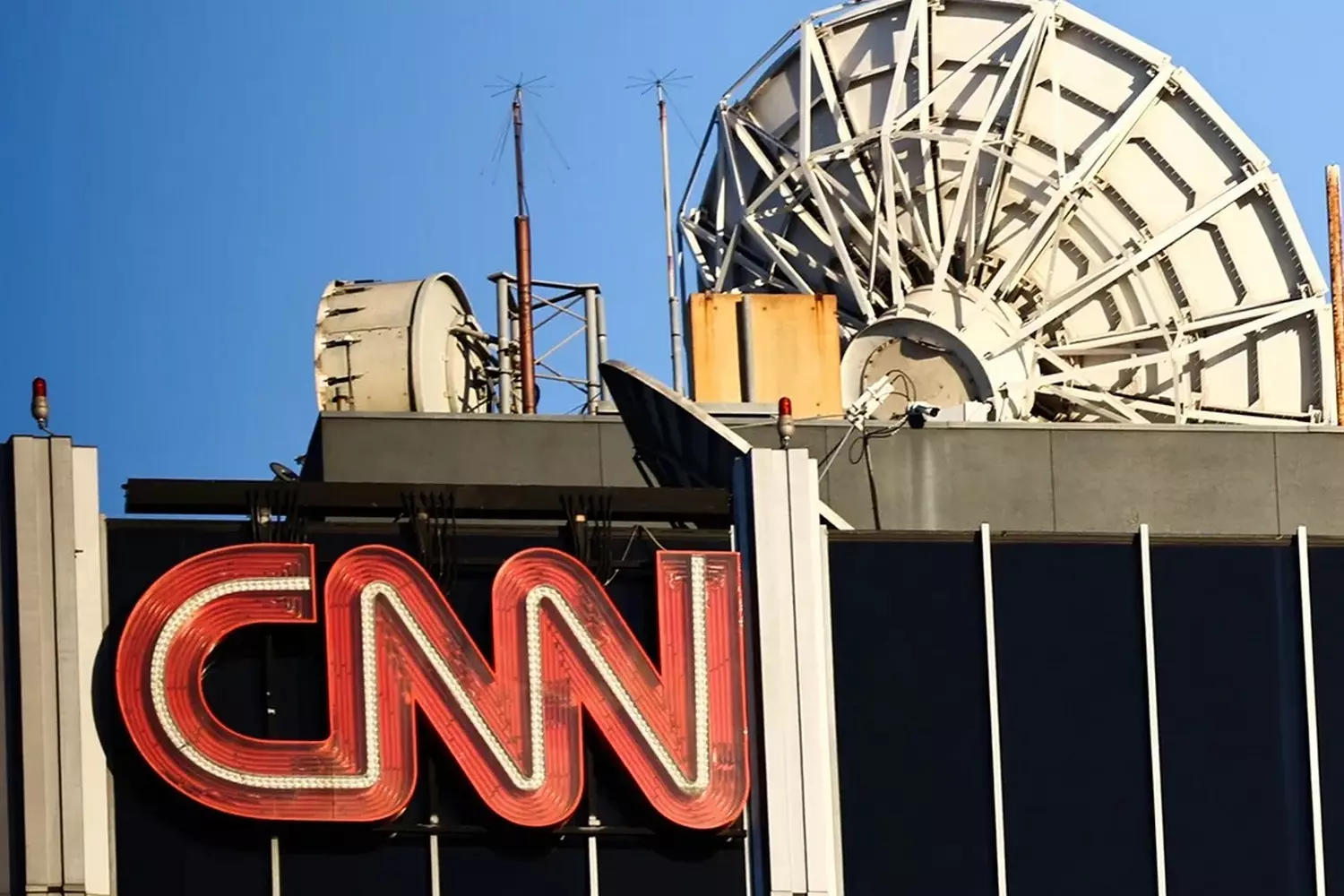 Вывеска CNN и телебашня: как новости попадают в эфир Узнаваемая вывеска CNN на здании с высокой телевизионной антенной на заднем плане, олицетворяющая мировое вещание