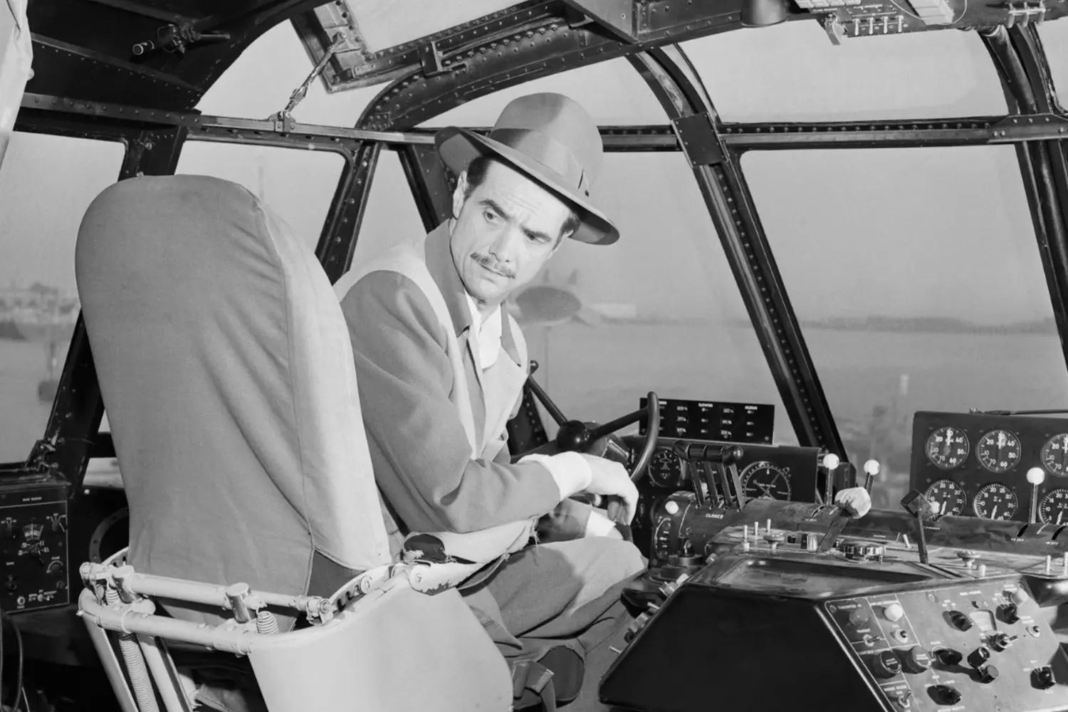 Howard Hughes in the Cockpit: The Aviator at His Natural Habitat Black-and-white photo of Howard Hughes sitting in the cockpit of his aircraft, hands on controls, with intense focus — capturing his legendary passion for aviation and engineering
