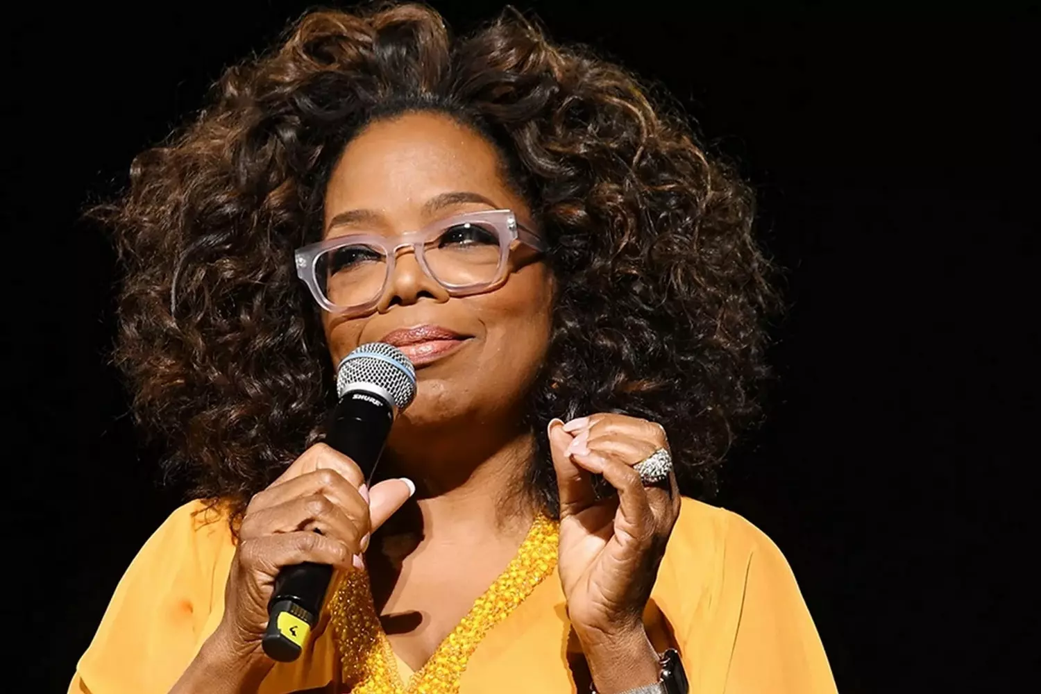 Опра Уинфри: Икона стиля, влияния и вдохновения Опра Уинфри уверенно улыбается в элегантном наряде, выступая с микрофоном на мероприятии