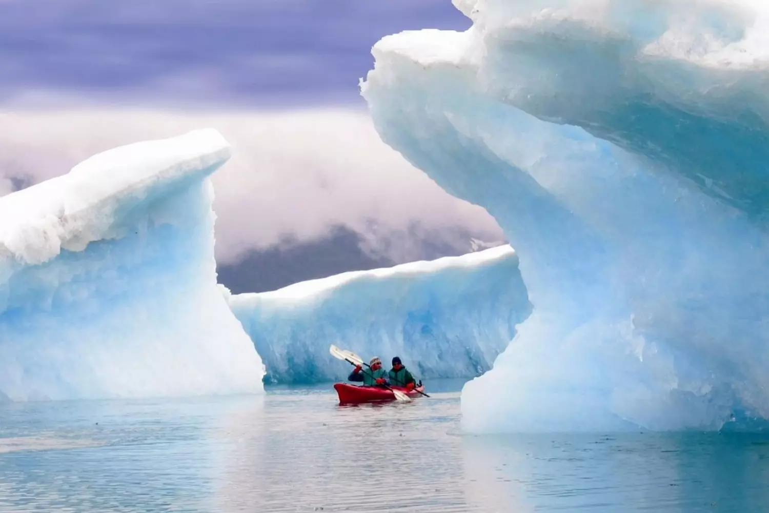 Paddling Through Ice Ages: Kayaking Alaska's Glacial Wonderland A lone red kayak glides between towering blue icebergs in Glacier Bay, with the paddler's reflection perfectly mirrored in the still, icy waters beneath Alaska's rugged coastal mountains