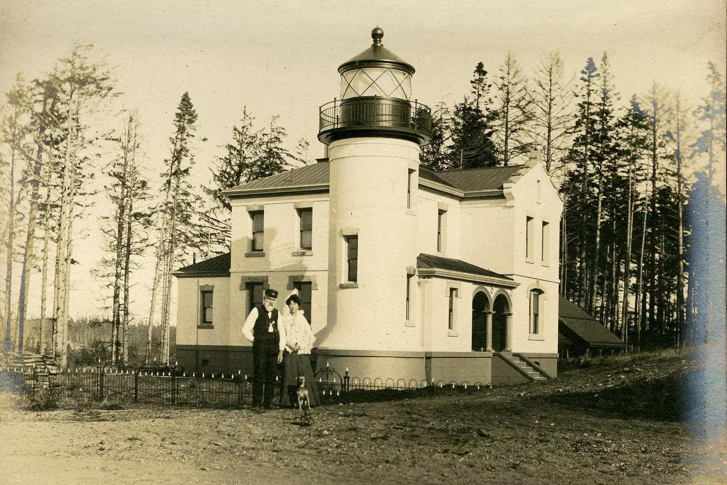 Historic photo of a lighthouse keeper, his wife, and a dog in front of the lighthouse