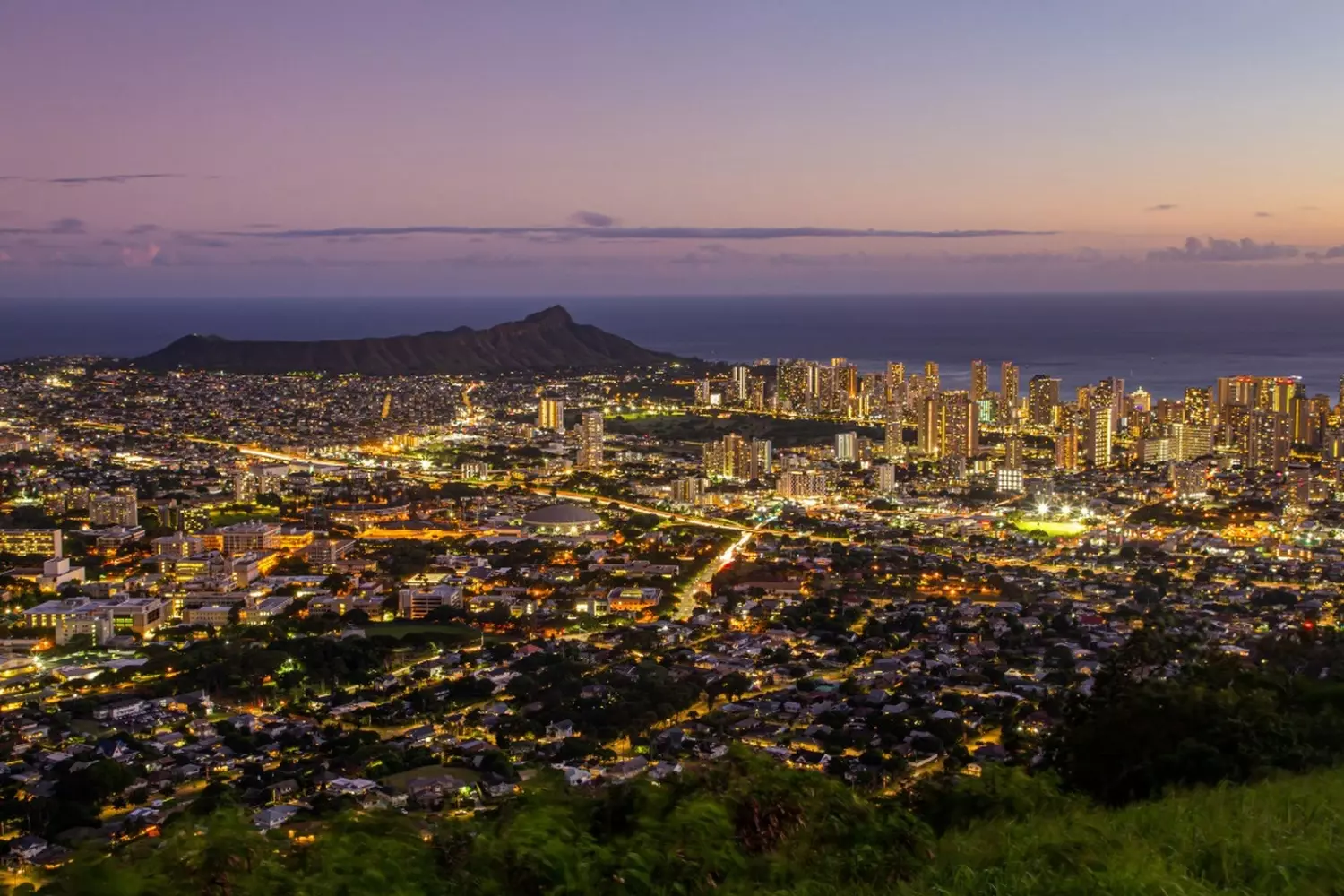 Hawaiian Nightscape: City Lights Meets Tropical Wilderness Beautiful long-exposure photograph capturing the contrast between the bright lights of a Hawaiian city and the dark, lush tropical mountains under a moonlit sky