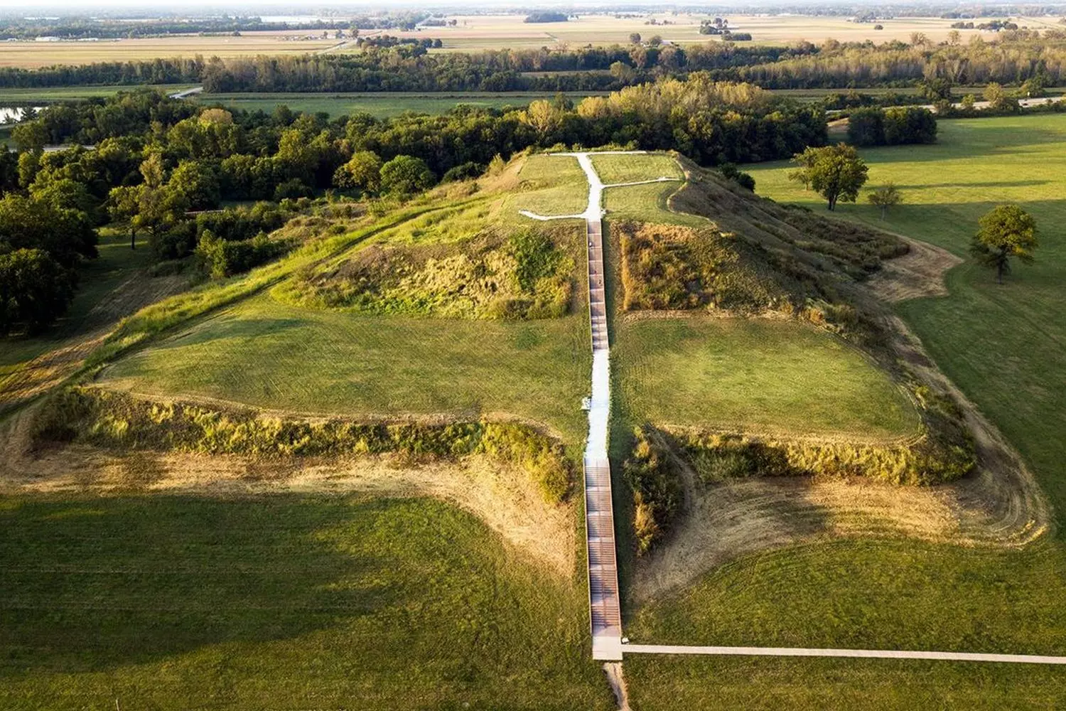 Echoes of Ancient Illinois: Exploring the Mysterious Indian Mounds Ancient Native American burial mound in Illinois surrounded by lush greenery under dramatic cloudy skies, with informational plaque visible in foreground showing its historical significance