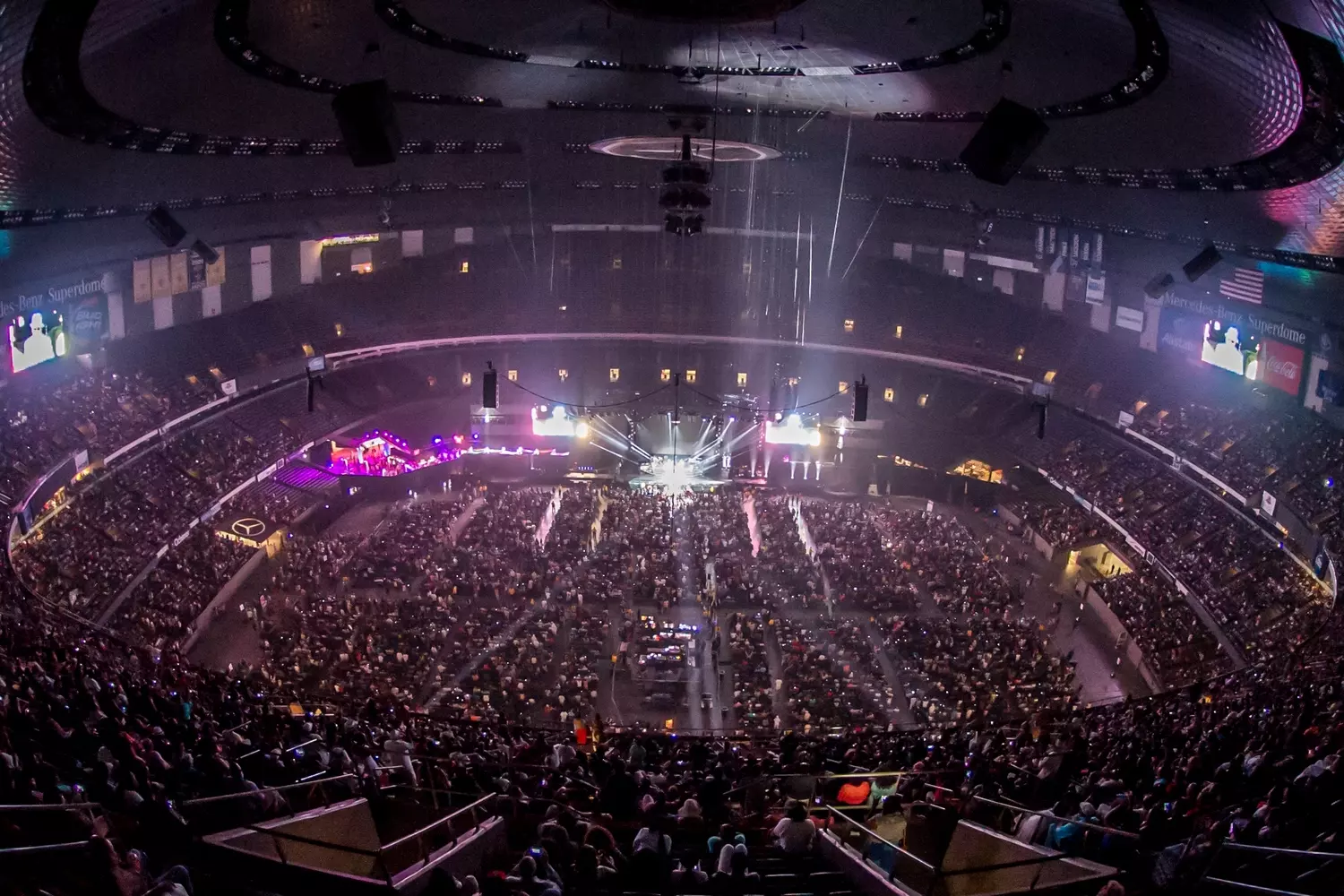 Огромный зал Mercedes-Benz Superdome во время Essence Festival, полностью заполненный людьми, сцена в центре, разноцветное освещение, масштабное шоу